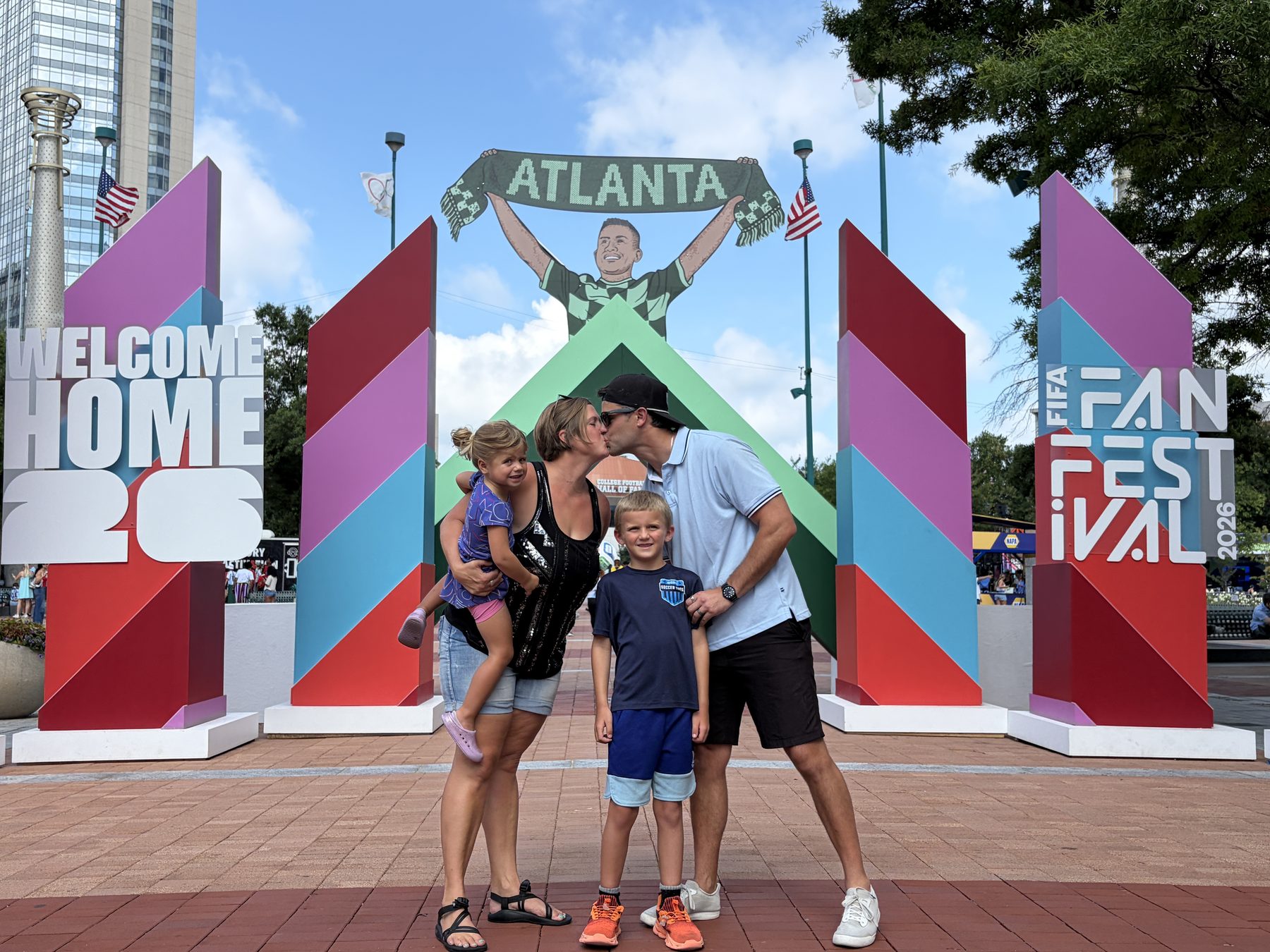 Josh, his wife, and their two kids kissing in front of a "Welcome Home ATL" FIFA Fan Festival display in downtown Atlanta.